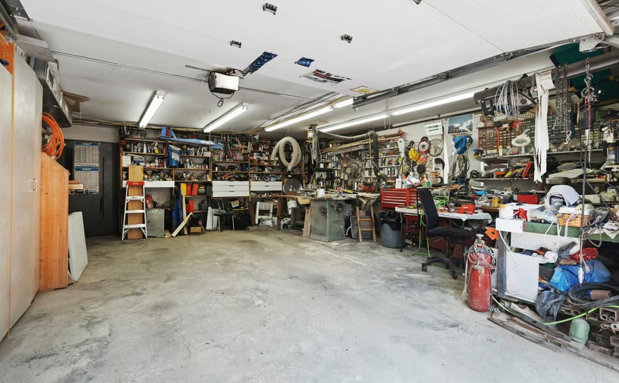 A domestic garage with bare concrete underfoot, shelving and stored equipment along the walls